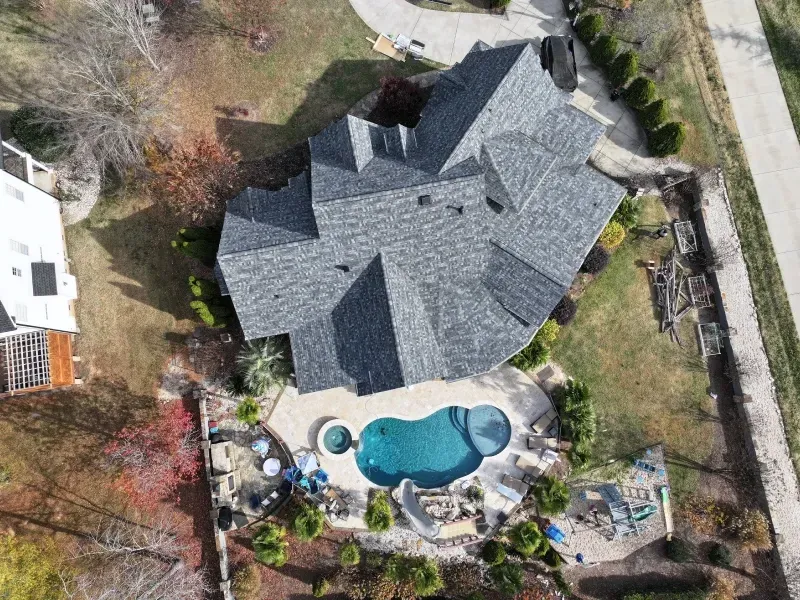 Aerial view of a house with a gray roof, backyard pool, patio seating, and surrounding trees.