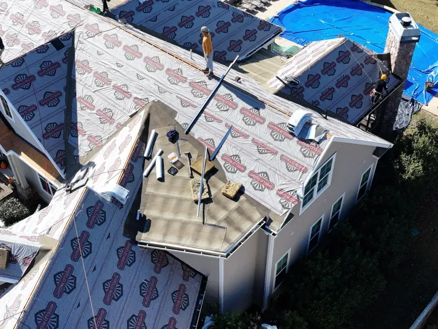 Aerial view of a house roof with damaged shingles, tarps, and several rooftop vents after storm repairs