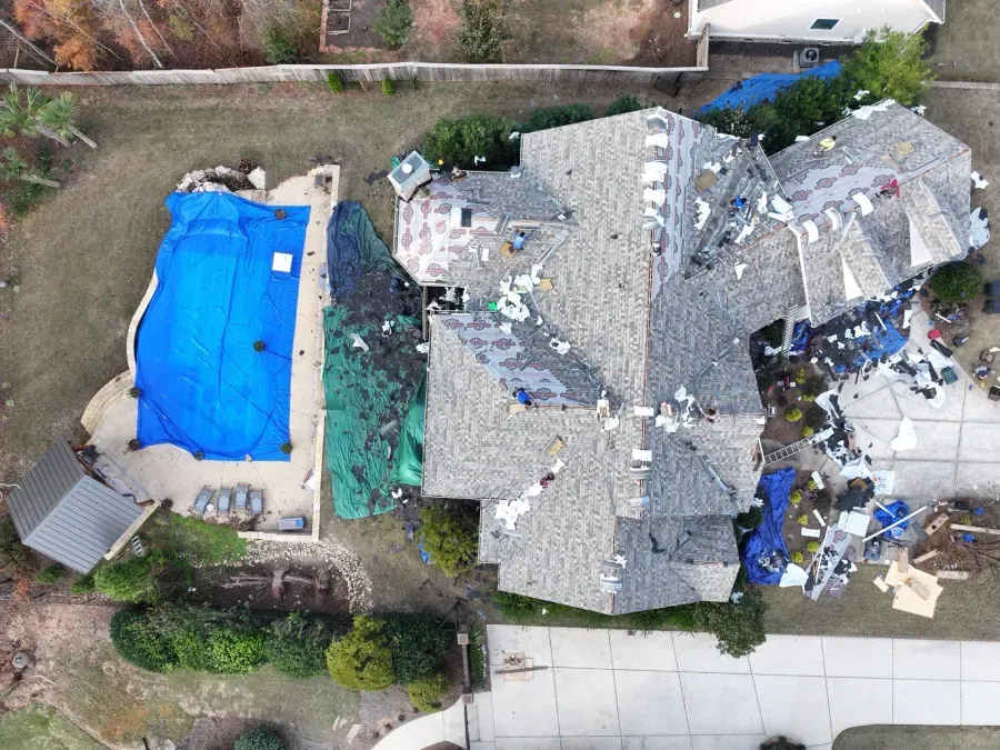 Aerial view of a house with storm-damaged roof beside a covered blue backyard pool and debris.