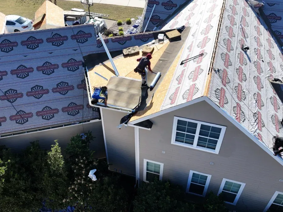 Workers installing shingles on a house roof, with underlayment exposed and patterned roofing visible nearby