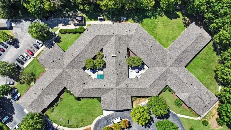 Aerial view of a gray-roofed building complex surrounded by green trees and parked cars.