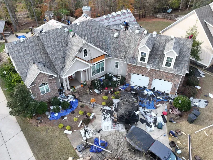 Aerial view of a house with roof damage and yard covered in debris and tarps.