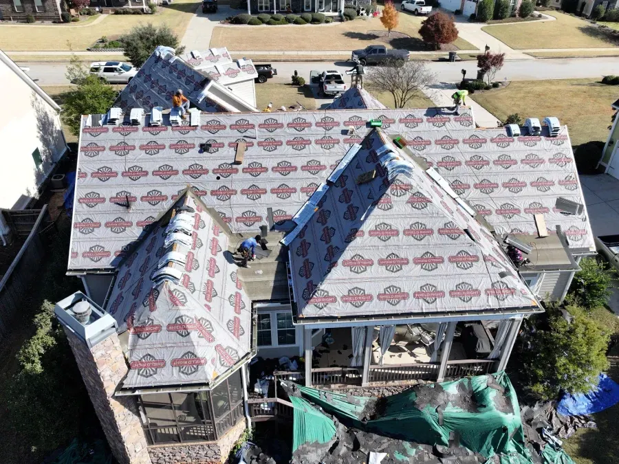 Aerial view of a house roof under repair, with workers and exposed sections covered in protective sheeting.