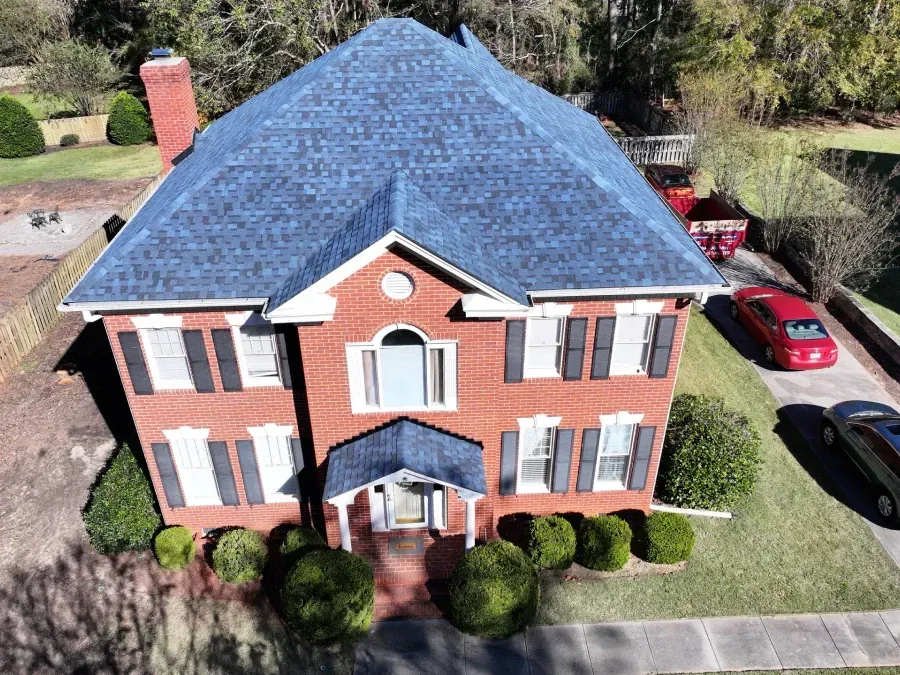 Aerial view of a red brick house with a blue roof, front lawn, and a red car parked nearby.