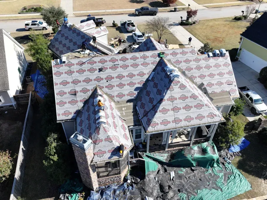 Aerial view of a house under roof repair, with exposed plywood, tarps, and construction materials around it