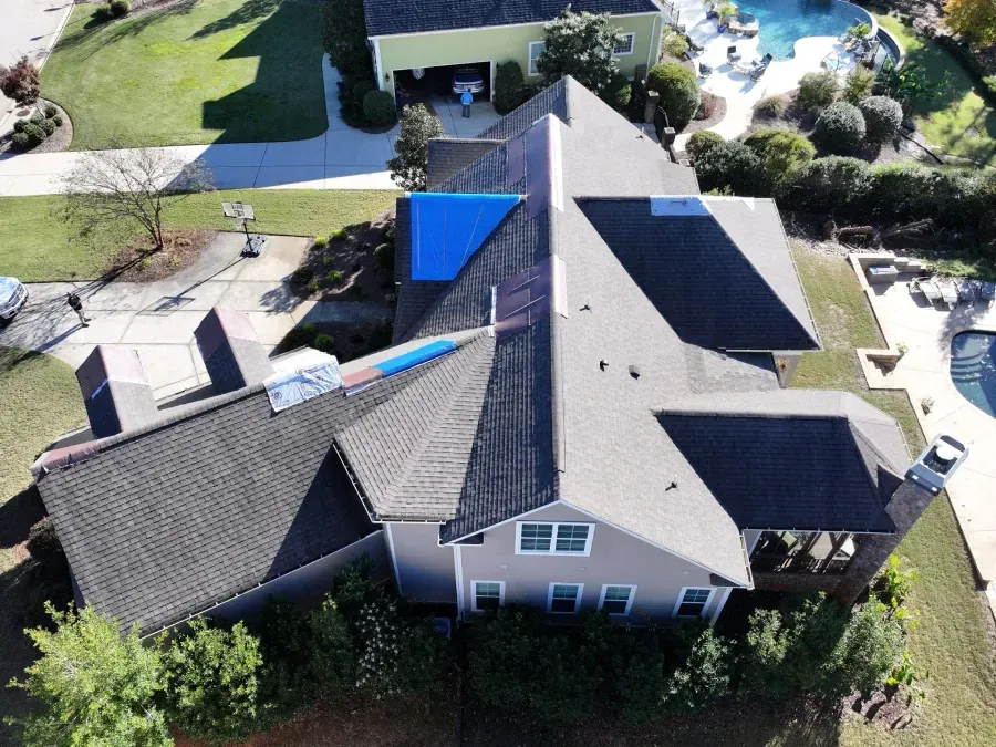 Aerial view of a house with a gray roof, blue tarp, and surrounding trees and yard