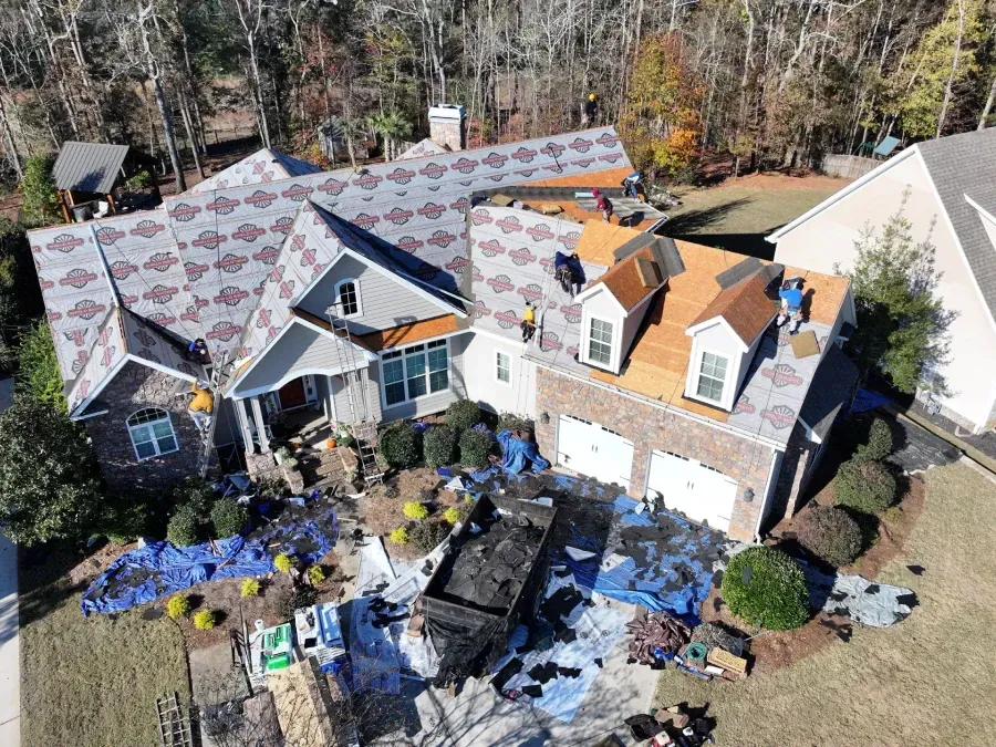House under roof repair with exposed plywood, blue tarps, and debris in front yard