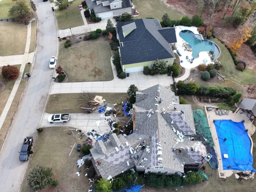 Aerial view of a storm-damaged house with roof debris, surrounded by neighboring homes and a backyard pool.