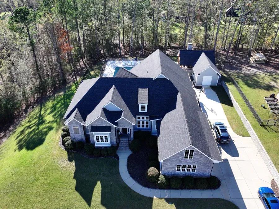 Aerial view of a large gray house with dark roof, driveway, and wooded yard
