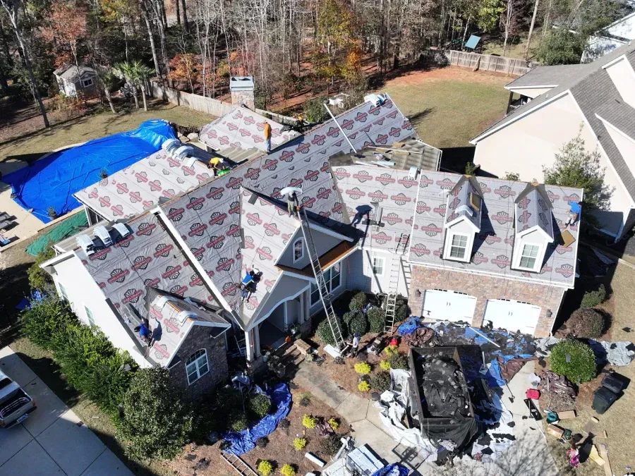 Aerial view of a house with a roof under repair, tools and debris in the driveway, and a blue pool nearby