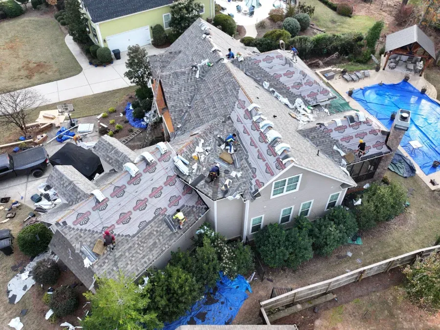Aerial view of a house with roof damage, debris, and blue tarps in the yard after a storm