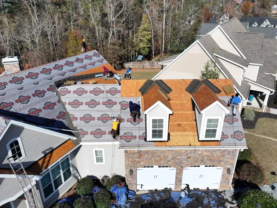 Damaged roof with missing shingles on a suburban house, with roofing materials and workers nearby