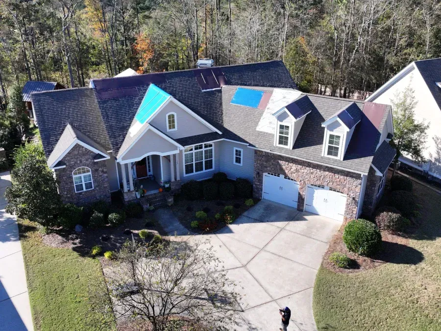 Aerial view of a gray suburban house with a dark roof, white garage doors, and a large driveway.