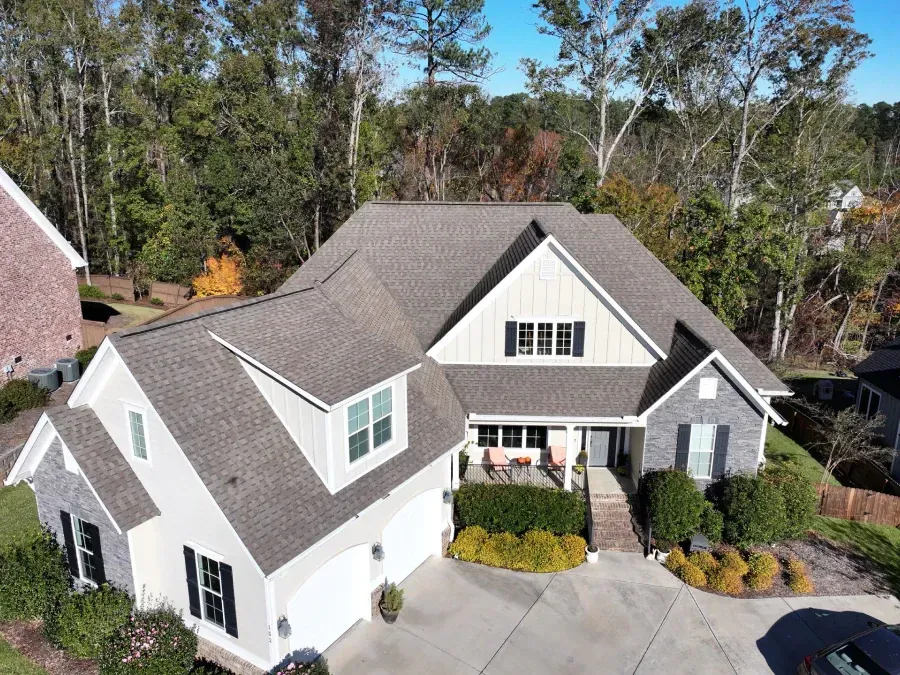 Aerial view of a gray and white suburban house with a landscaped front yard and trees behind it