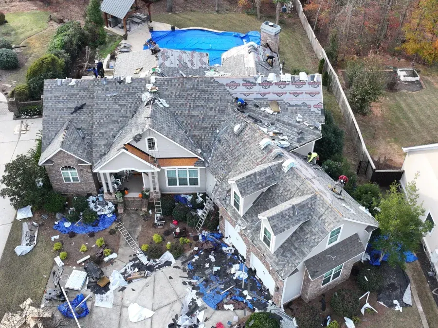 Damaged house roof covered in debris after storm, with scattered patio furniture and a blue tarp in the yard
