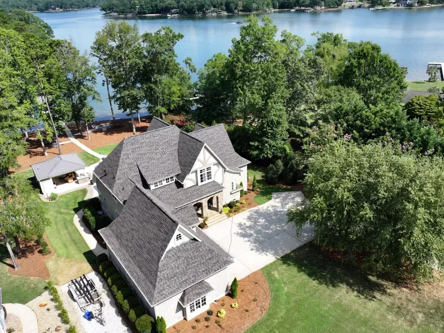 Aerial view of a gray house with a driveway, trees, and a lake in the background