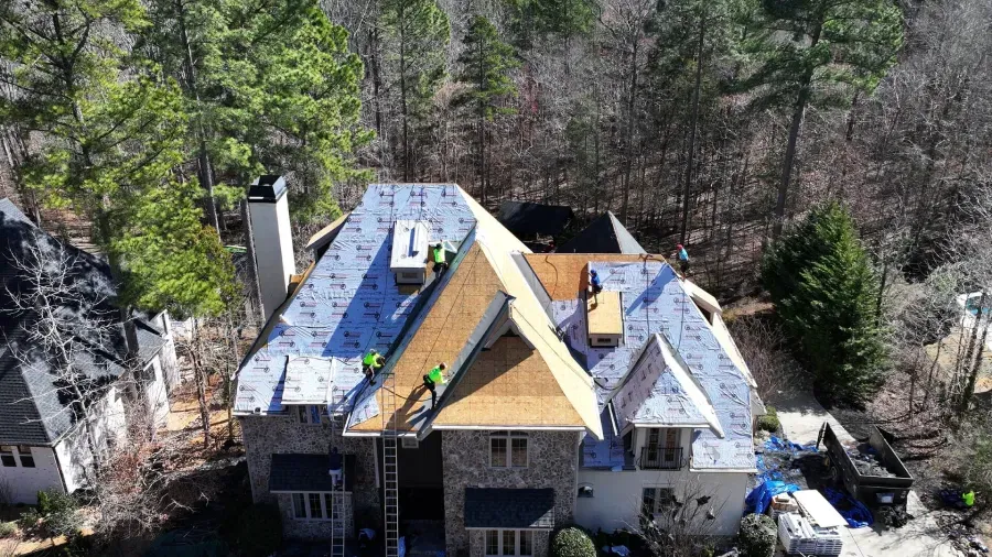 A house roof under repair with blue underlayment, exposed plywood, and a ladder in front.