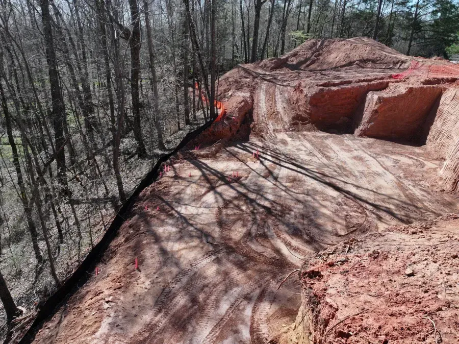 Wide red clay slope beside leafless forest, with carved cutouts and shadows across the ground.
