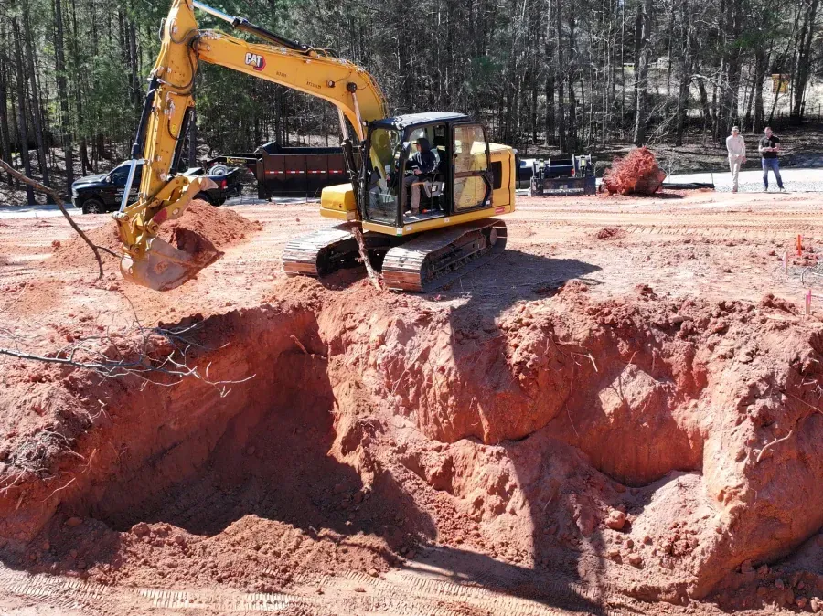Excavator digging a large red dirt pit at a construction site with trees in the background