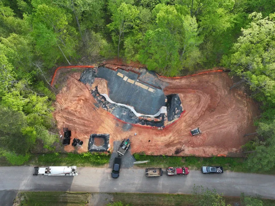 Aerial view of a house under construction surrounded by red dirt, trees, and parked vehicles.