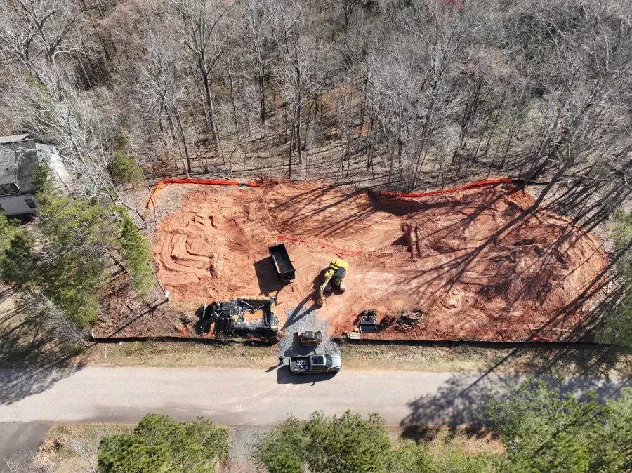 Construction site with excavator and dump truck on orange dirt beside a road and wooded area