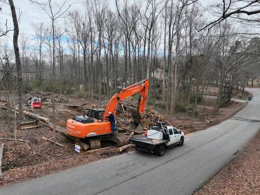 Orange excavator demolishing a white pickup truck on a roadside in a wooded area
