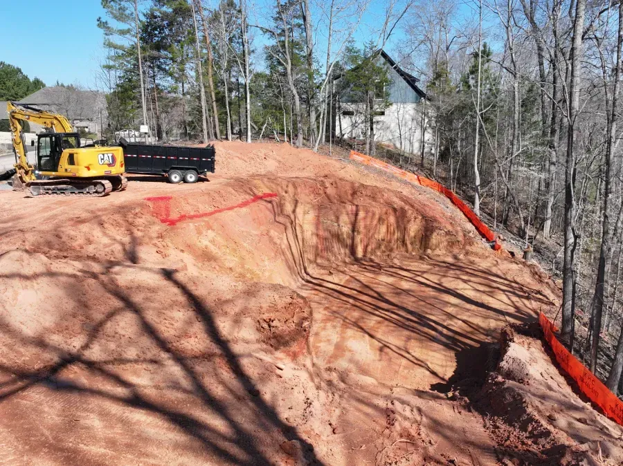 Excavator and dump truck on a red dirt hillside construction site with trees in the background