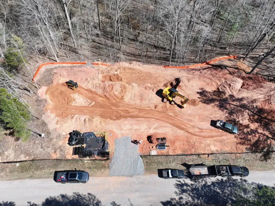 Aerial view of a dirt construction site with excavator, parked cars, and bare trees around it