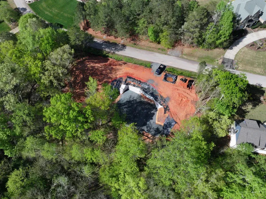 Aerial view of a house under construction surrounded by trees and red dirt, with vehicles on a nearby road