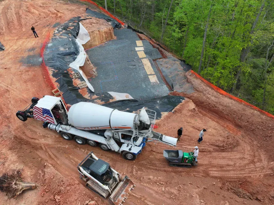 Concrete truck and workers at a road construction site on a dirt slope with orange safety fencing.