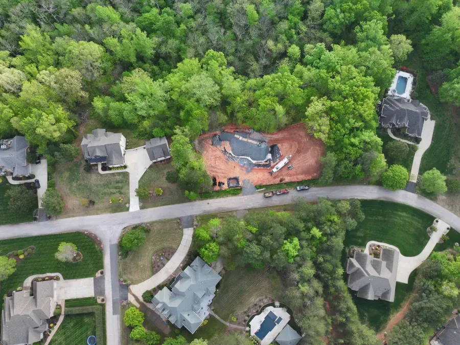 Aerial view of houses along a curving road beside dense green trees and a red dirt construction site