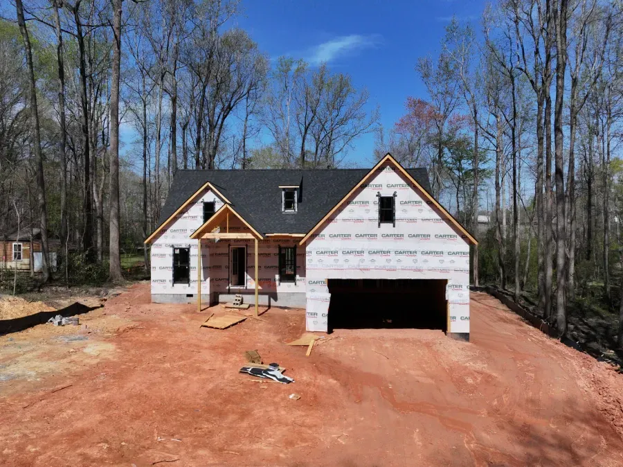 New house under construction with white siding, dark roof, and unfinished red dirt yard