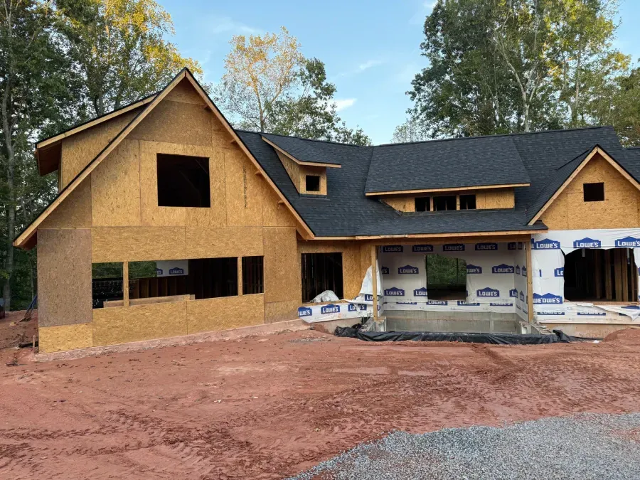 Unfinished house with wood siding and dark roof under construction on a red dirt lot