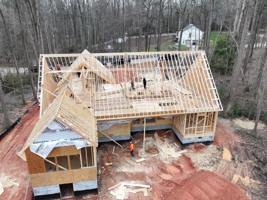 Aerial view of a house under construction with exposed wooden framing and a partially covered roof in a wooded area