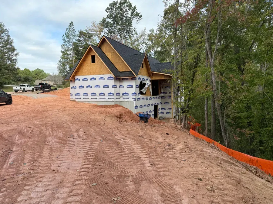 Partially built house wrapped in white house wrap on a red dirt lot beside trees and orange safety fencing