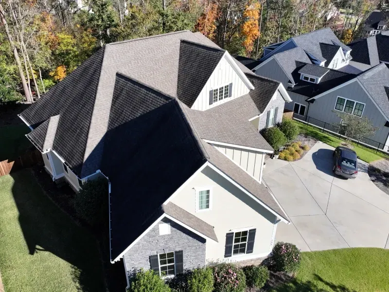 Aerial view of a suburban house with a gray roof and driveway, surrounded by autumn trees.
