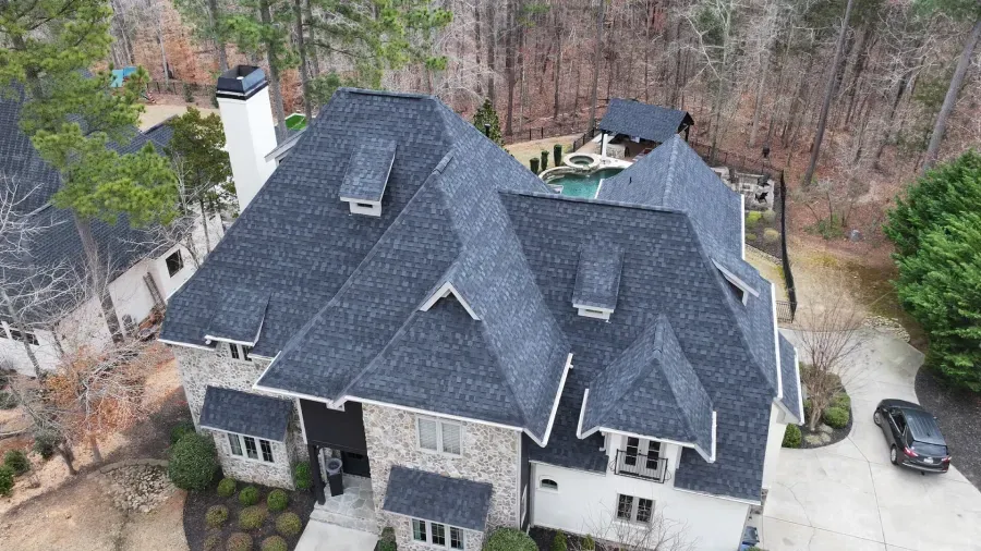 Aerial view of a large gray house with a dark roof and driveway beside wooded trees.