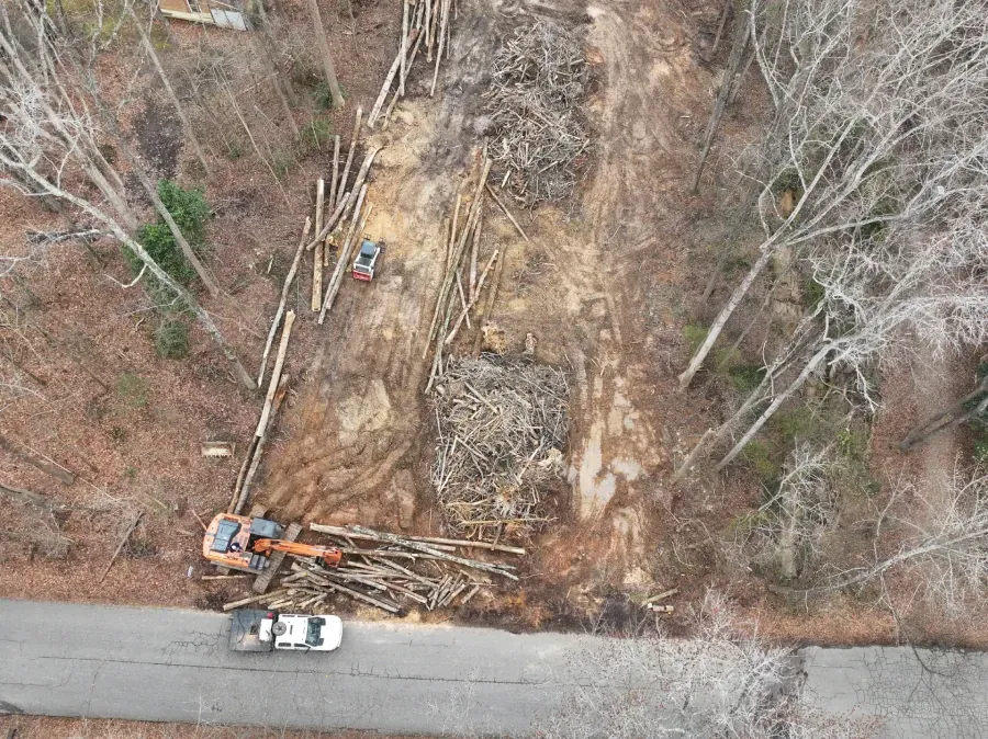 Aerial view of roadside excavation with a truck, logs, and heavy equipment clearing a wooded lot