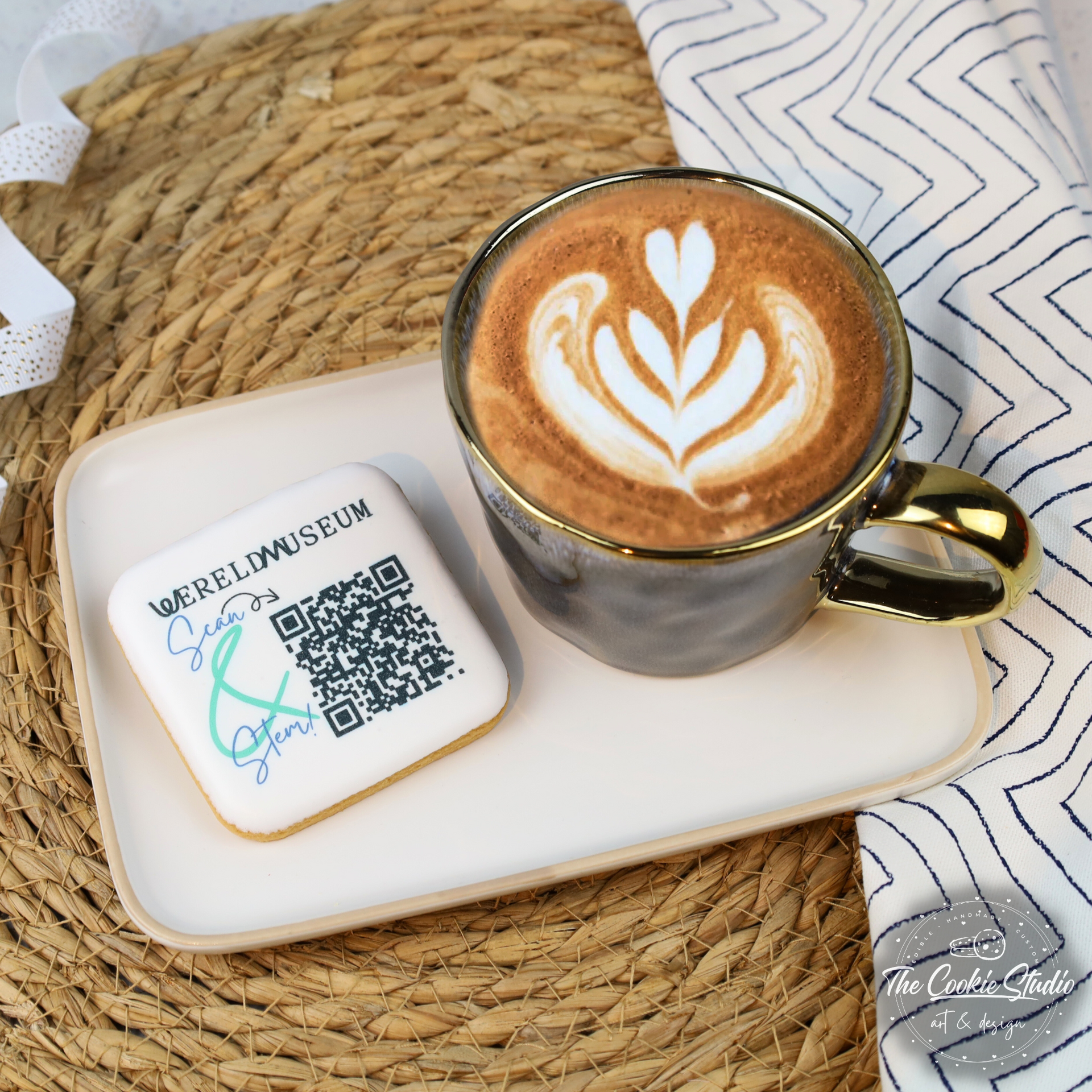 Een latte met latte art en een koekje met een QR-code, geserveerd op een wit bord.
