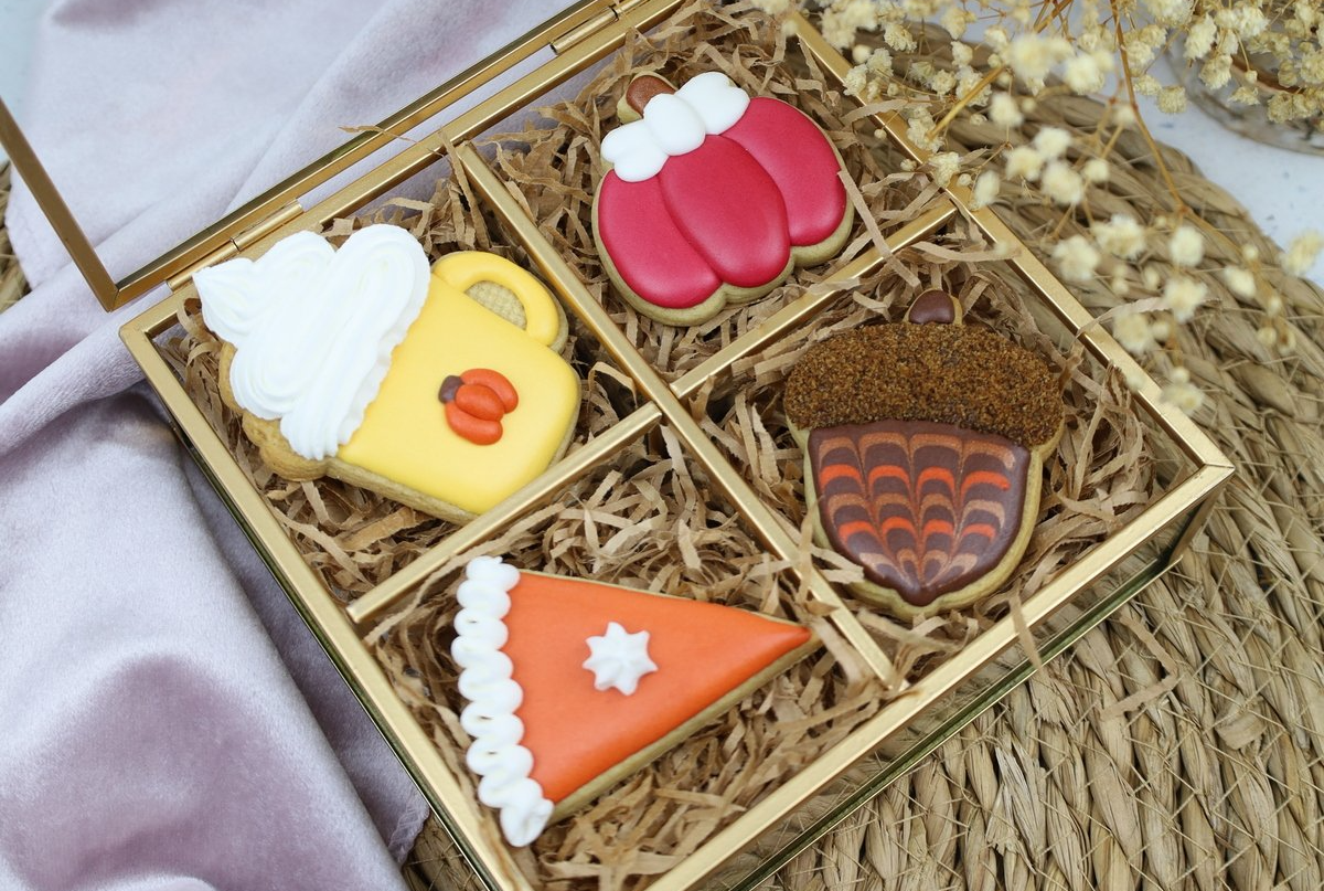 Vier suikerkoekjes met herfstthema en glazuur, verpakt in een gouden glazen doos. De koekjes hebben de vorm van een mok, wanten, een eikel en een taart.