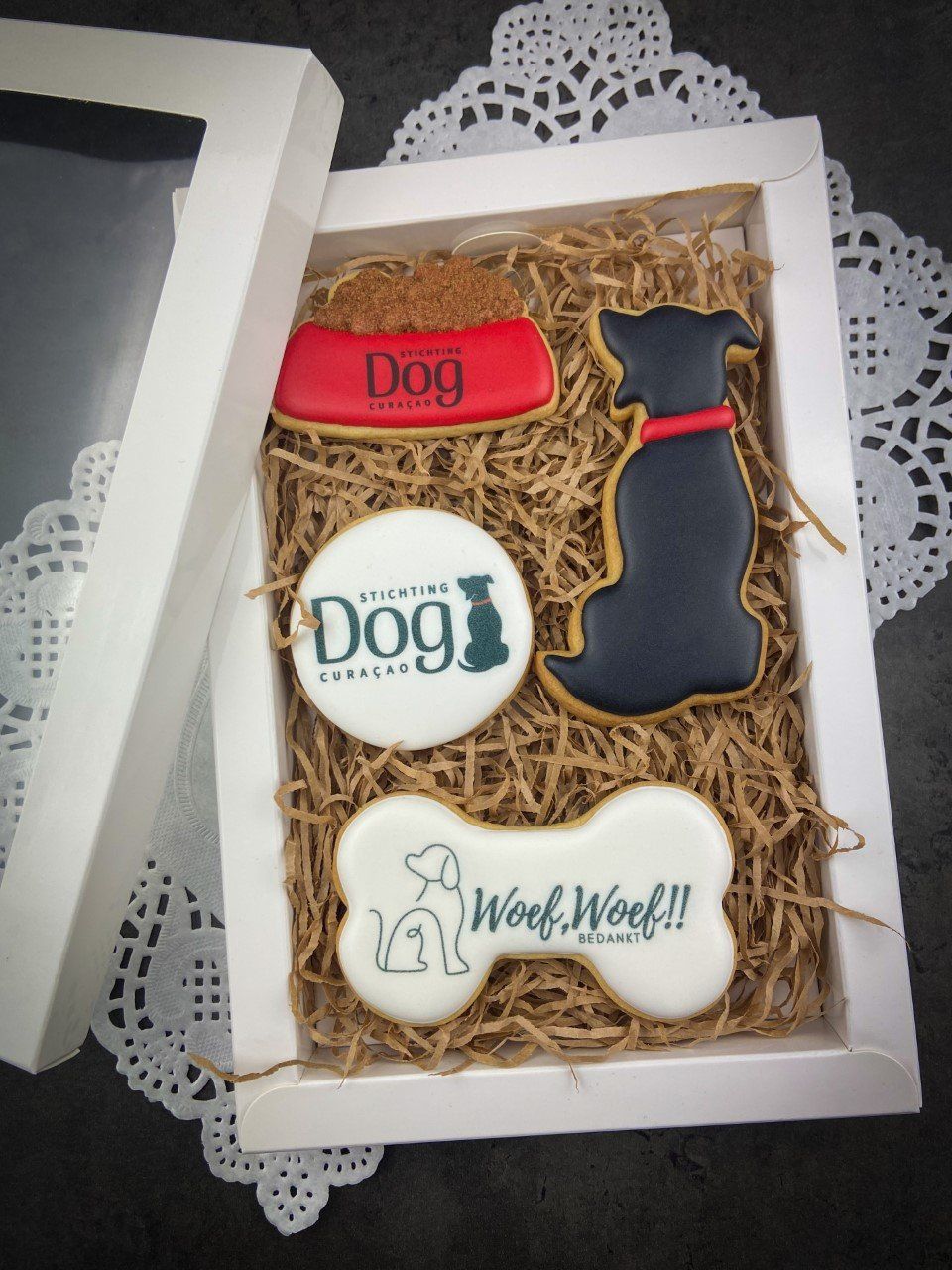 Doos met versierde koekjes met hondenthema: koekjes in de vorm van een hondenkom, een hondensilhouet, een bot en een logo, op een rietje in een witte doos.