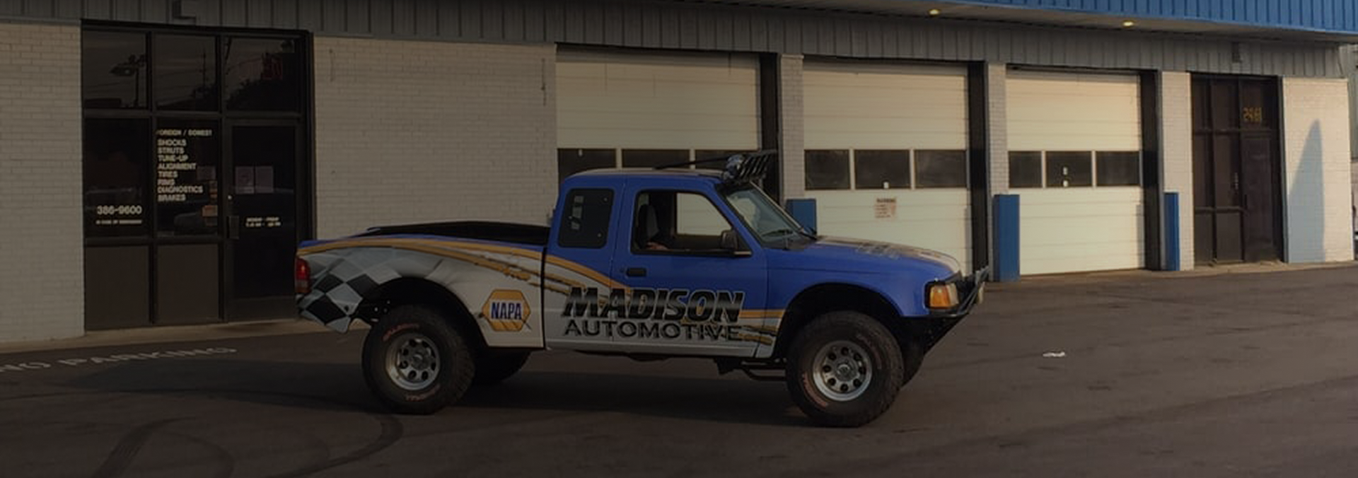 A blue and white racing truck parked in front of a garage with closed doors. | Madison Automotive
