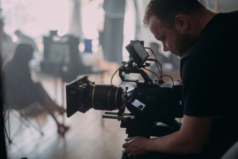Una persona manovra una telecamera professionale sul set di uno studio, con un soggetto sfocato sullo sfondo.