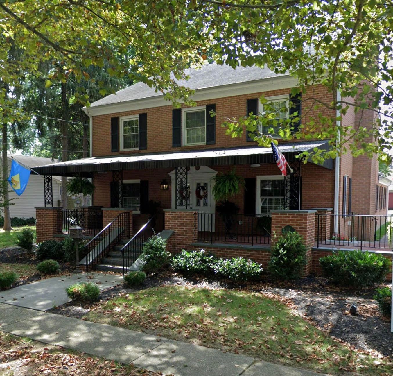 Brick two-story house with black shutters, covered porch, and American flag. Green bushes and trees surround the building.