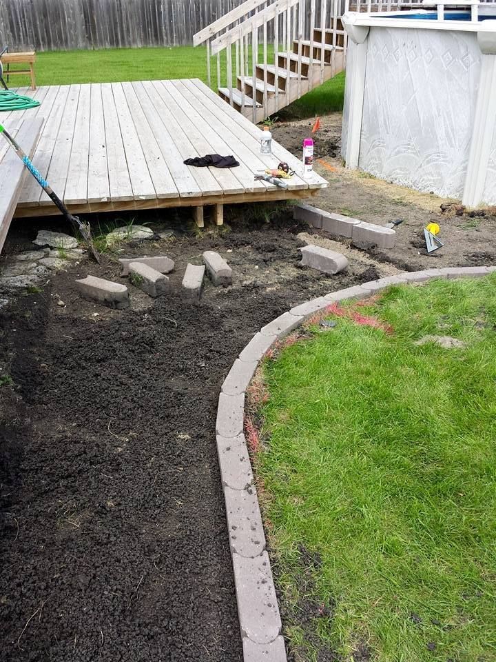 Backyard with a deck, pool, and path being built. Dark soil, gray bricks, green grass.