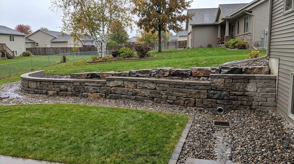 Stone retaining wall and terraced lawn in a suburban backyard, with a gravel path and houses in the background