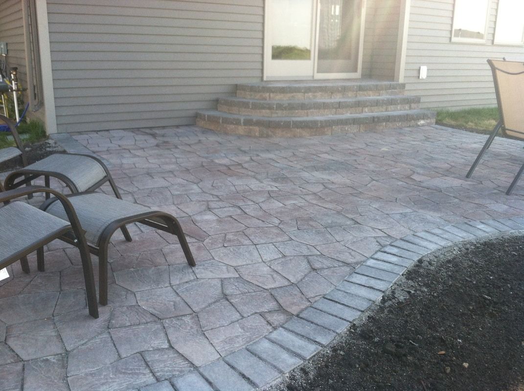 Patio paved with interlocking stones, bordered by dark pavers, next to a house with steps leading to a door;