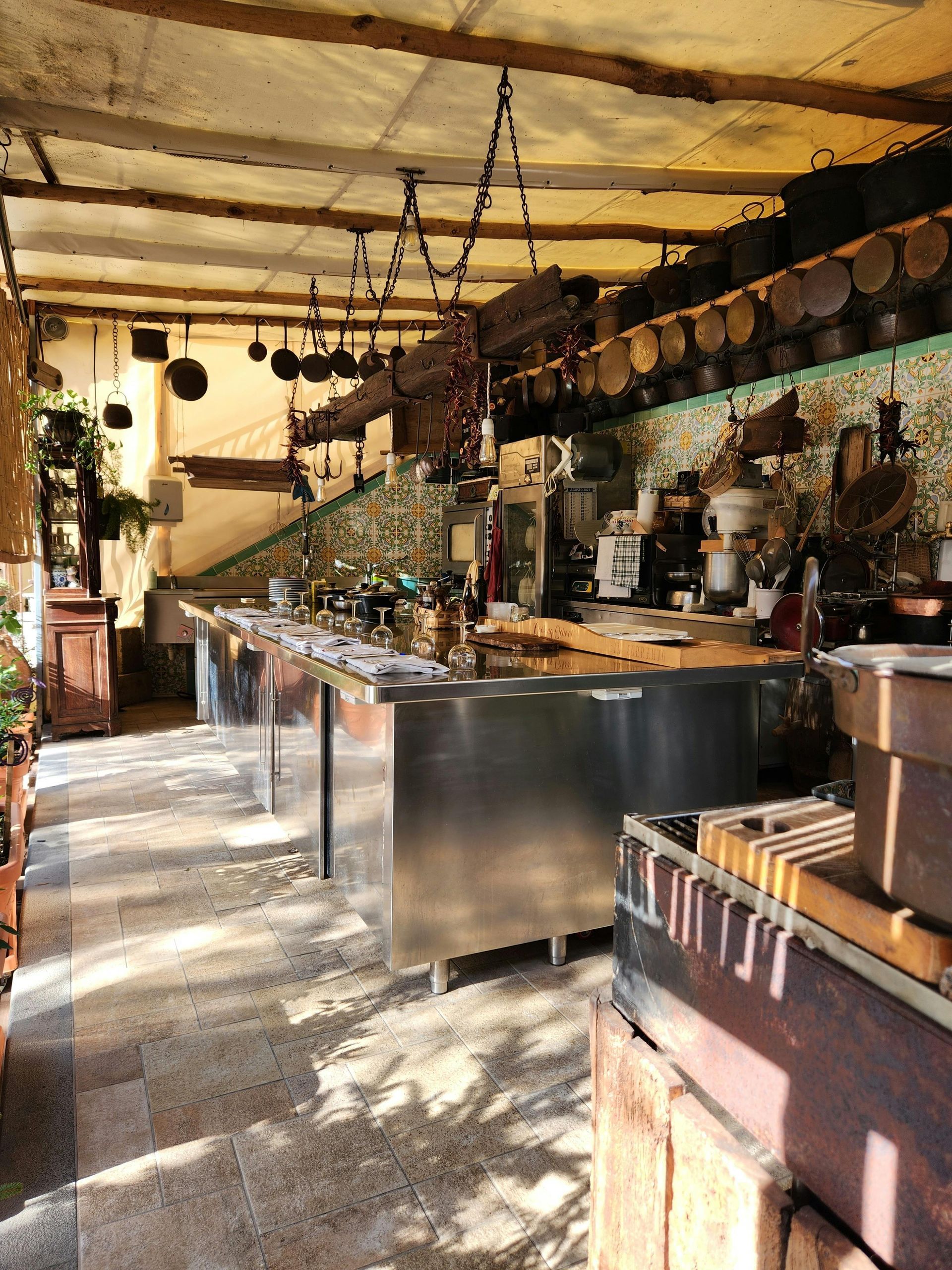Rustic outdoor kitchen with stainless counters, hanging pans, and dappled sunlight under a shaded roof
