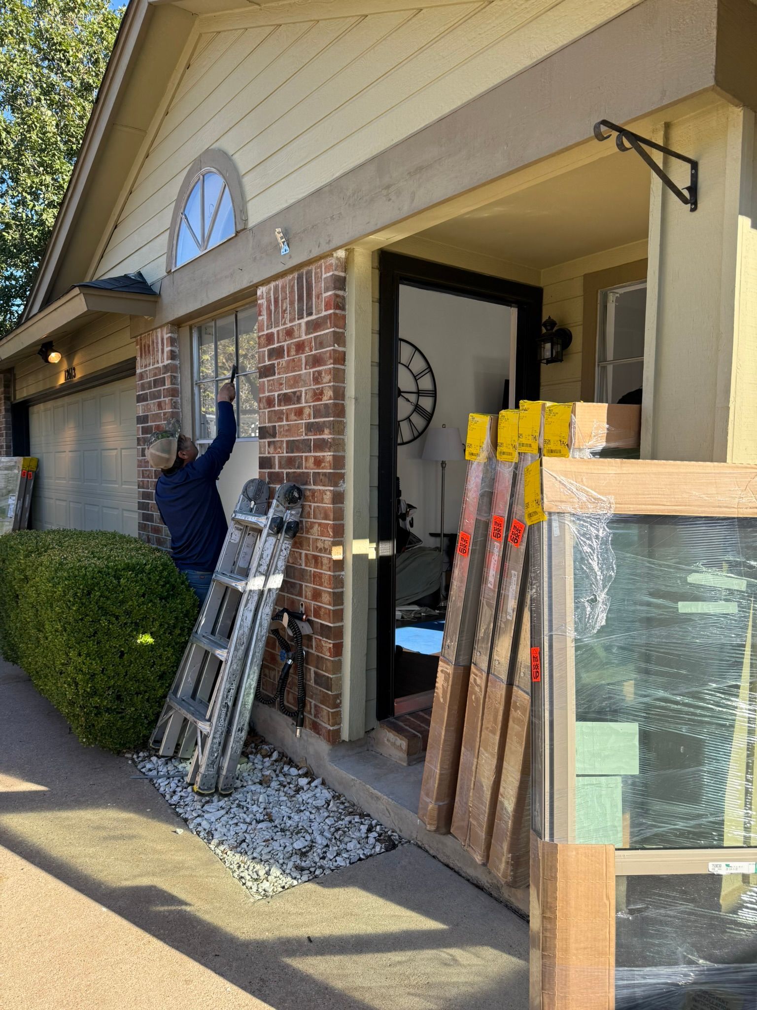 Man on ladder outside a home working on a window, brick facade. Lumber and a window frame are present.