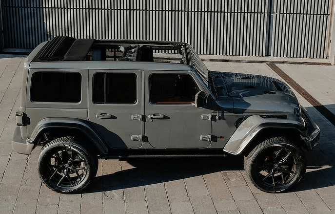 A gray jeep with a sunroof is parked on a sidewalk in front of a building.